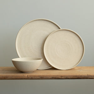 Tableware set with a large and small upright plate with a straight edge and a breakfast bowl in front of it in matt beige with fine sprinkles on a wooden table