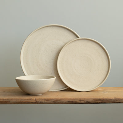 Tableware set with a large and small upright plate with a straight edge and a breakfast bowl in front of it in matt beige with fine sprinkles on a wooden table