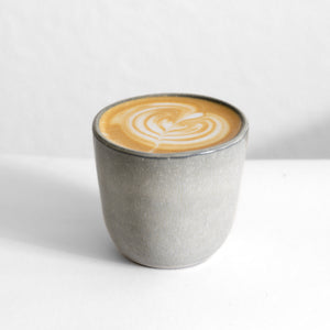 Hellgrauer Cappuccino Becher ohne Henkel gefüllt mit einem Kaffee mit Latte Art