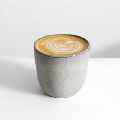 Hellgrauer Cappuccino Becher ohne Henkel gefüllt mit einem Kaffee mit Latte Art