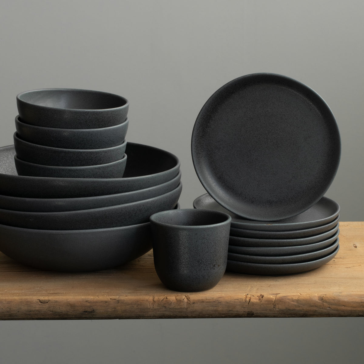 Matt black stoneware tableware arranged on a wooden board. On display are pasta plates, snack bowls, bread plates and a cappuccino cup