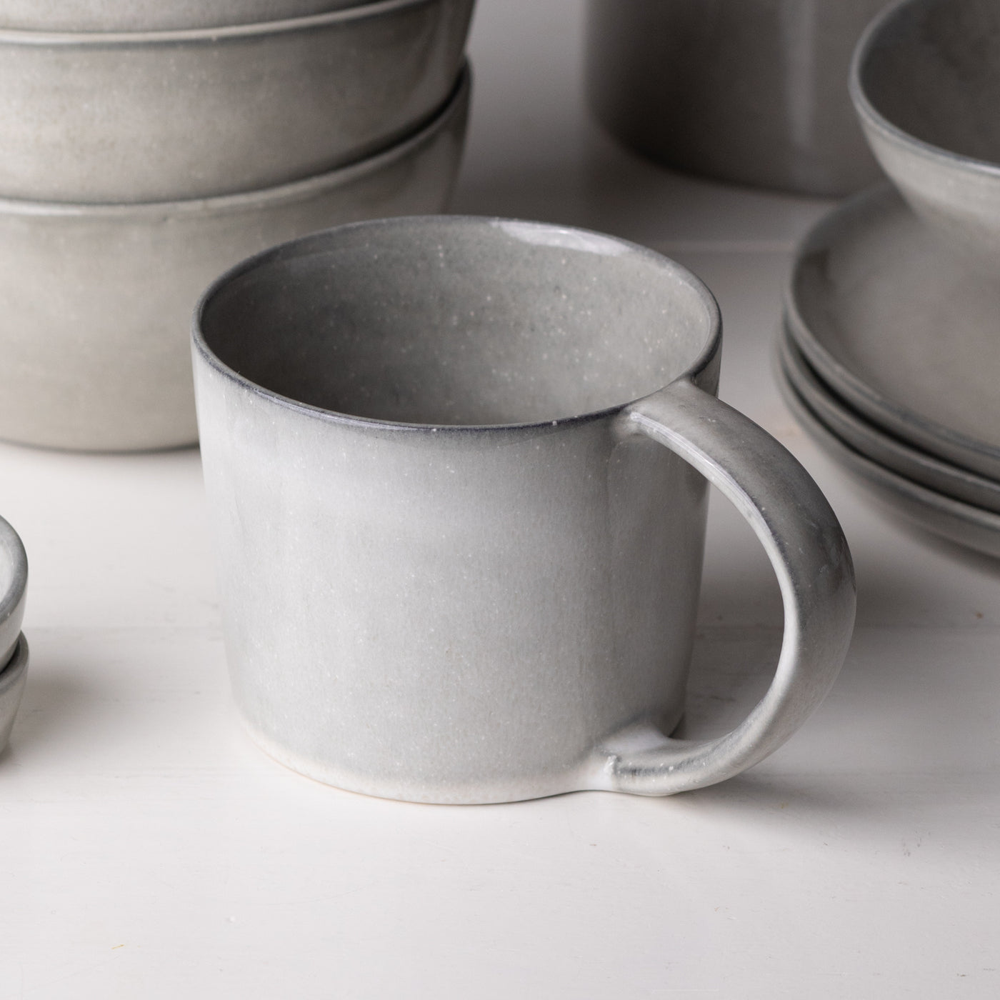 Im Vordergrund steht eine grosse Teetasse mit Henkel mit hellgrauer reaktive Glasur aus Steinzeug im Hintergrund sind grosse Teller und Müesli Bowls zu sehen