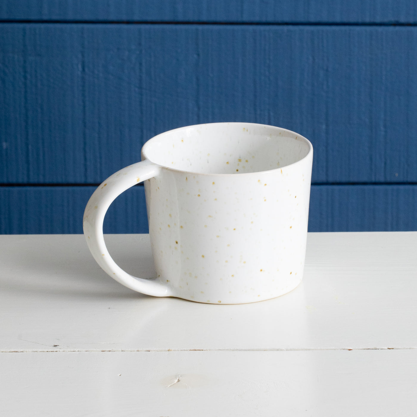 Large ceramic teacup with handle with white reactive glaze with golden brown spots in front of a blue wall