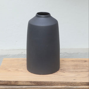 Le vase noir mat, avec une petite ouverture en haut et une large partie inférieure, est posé sur une planche en bois.